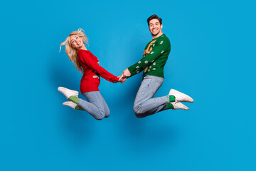 Fototapeta na wymiar Full length body size photo couple holding hands jumping up in ugly xmas sweater isolated bright blue color background