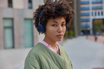 Photo of serious good looking woman with Afro hair listens music in headphones enjoys audio player dressed in casual jacket uses wireless headphones poses against blurred background outside.