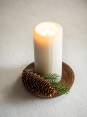 Minimalistic christmas composition, white candle on wooden stand with fir branch and pine cone on gray concrete background in morning light