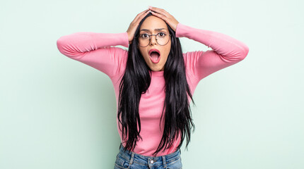 pretty hispanic woman feeling horrified and shocked, raising hands to head and panicking at a...
