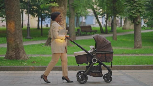 Busy female employee talking on phone, pushing stroller in park, maternity leave