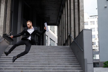 A cheerful and happy businessman in a suit holding a briefcase and a folder with documents, jumping...