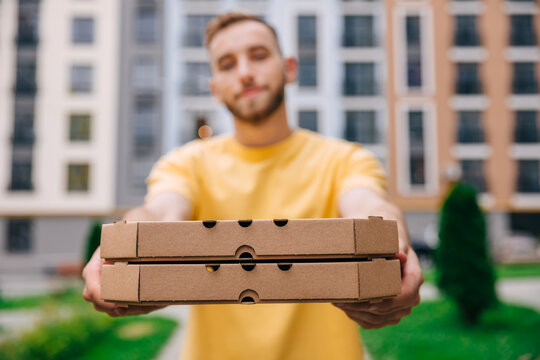 Shooting a portrait of a handsome delivery man standing in the street holding cardboard boxes while handing over the camera. Male courier handing pizza to customer.