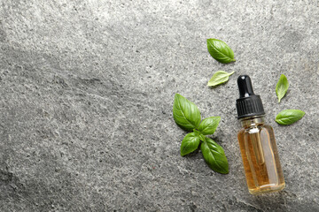 Glass bottle of basil essential oil and leaves on grey stone table, flat lay. Space for text