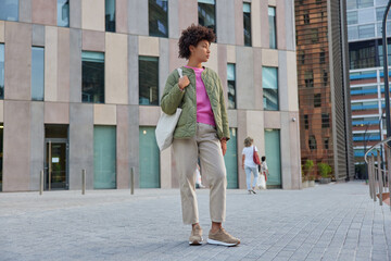 Full length shot of pensive young African American woman dressed in stylish clothes carries bag looks away walks in city passes by modern urban buildings finds out new places stands on pavement © WHstudio Leushin N