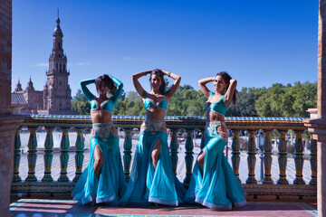 Three middle-aged Hispanic women, wearing turquoise dresses and rhinestones, to belly dance, posing next to a tiled railing. Belly dance concept.