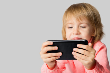 A child, a cute little girl dressed in a pink knitted sweater, holds a modern smartphone, playing and chatting on the Internet in social networks.