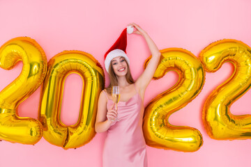 Smiling girl raises up glass of champagne in Santa's hat. golden air balloons 2022. New Year party 