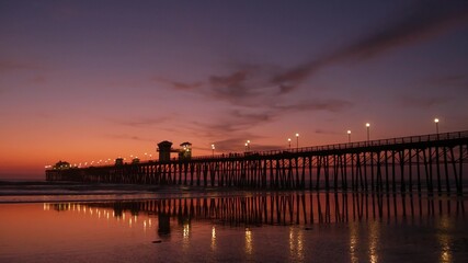 Fototapeta na wymiar Pier silhouette Oceanside California USA. Pacific ocean tide tropical beach. Summertime gloaming atmosphere. Purple aesthetic gradient, calm twilight sky, pink violet dusk. Lights reflection in water.