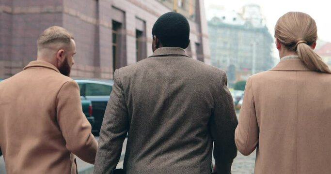 Multinational group of businessmen: two men and one woman walking down the street on way to office discussing new project. View from back.