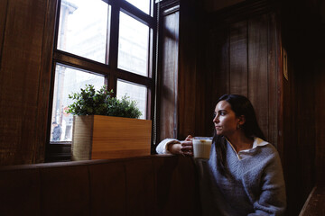 Lonely young woman drinking coffee and looking out the window in a wooden house