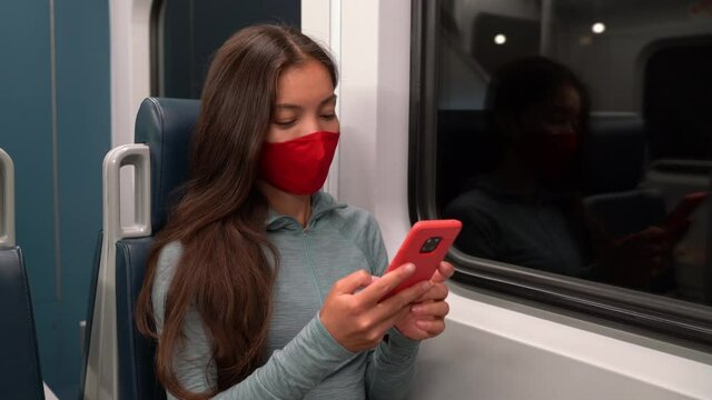 Woman wearing face mask in public transportation during coronavirus Covid-19 pandemic. Train transport commuter concept. Multiracial woman passenger using smart phone with face covering on commute