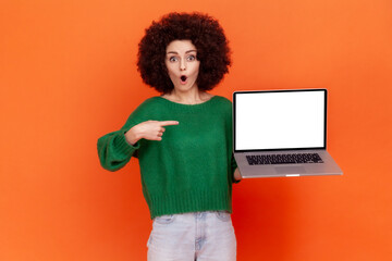 Obraz na płótnie Canvas Shocked woman with Afro hairstyle wearing green casual style sweater pointing at white empty display of her portable computer, copy space. Indoor studio shot isolated on orange background.