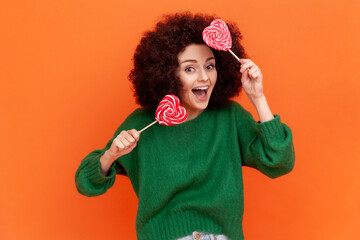Positive satisfied woman with Afro hairstyle wearing green casual style sweater holding two heart shape lollipops, having fun, funny expressions. Indoor studio shot isolated on orange background.