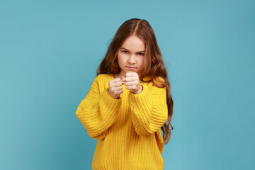 Angry little girl standing with raised fists, being ready to fight, looking with serious expression, wearing yellow casual style sweater. Indoor studio shot isolated on blue background.