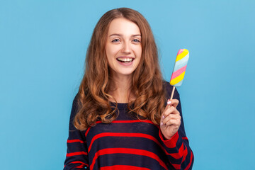 Satisfied woman wearing striped casual style sweater, holding rainbow ice cream, looking at camera with positive expression, feels hungry. Indoor studio shot isolated on blue background.