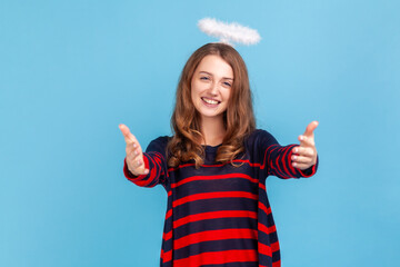 Cute woman wearing striped casual style sweater and nimb over head, raising hands in gesture welcome, come into my arms, sharing free hugs. Indoor studio shot isolated on blue background.
