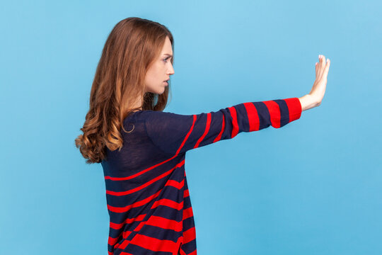 Side View Of Scared Serious Young Woman Wearing Striped Casual Style Sweater Showing Block Stop Gesture, Set Back Bullying And Violence. Indoor Studio Shot Isolated On Blue Background.