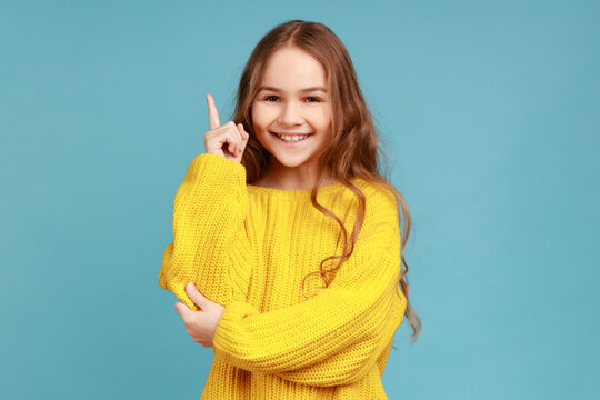 Portrait of little girl points finger up, looking inspired by genius thought, showing good idea sign, wearing yellow casual style sweater. Indoor studio shot isolated on blue background.