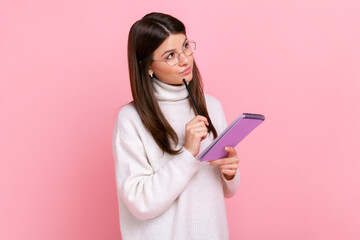 Pensive brunette female writing in paper notebook, making to do list or plan, looks away thoughtfully, wearing white casual style sweater. Indoor studio shot isolated on pink background.