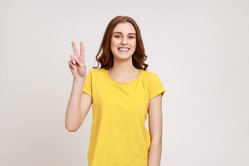 Beautiful satisfied teenager girl showing v sign symbol of peace with fingers, looking at camera with happy smile, celebrating triumph. Indoor studio shot isolated on gray background.