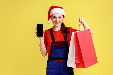 Delivery woman shows smart phone with blank display, holds shopping bags with goods odered online, wearing blue overalls and santa claus hat. Indoor studio shot isolated on yellow background.