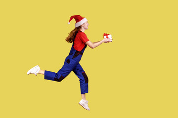 Fototapeta na wymiar Side view full portrait of courier female running with gift box to deliver Christmas for clients, wearing blue overalls and santa claus hat. Indoor studio shot isolated on yellow background.