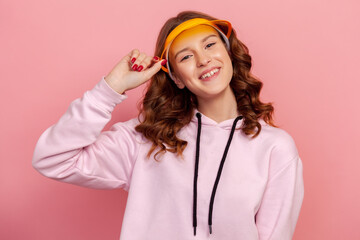 Portrait of happy curly haired teenage girl in hoodie touching yellow sun visor cap and sincerely...