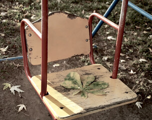 an unpaired children's swing with a yellow back with iron red handrails an autumn leaf lies on the seat