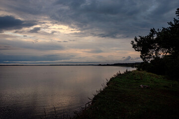 Fototapeta na wymiar coastal landscape of big calm lake in Latvia, green grass, cloudy dramatic sky, evening sunset light in blue hour