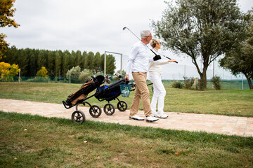 Senior couple in elegant clothes and with golf clubs walking together to the course to play golf.