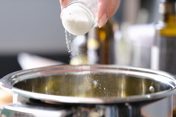Woman hand pours fine salt into saucepan closeup