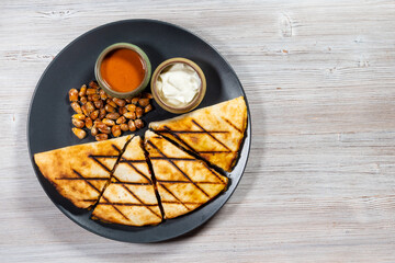 top view of cut Quesadilla from folded tortilla filled with cheese, meats, spices with sauces and fried corn seeds on plate on wooden board with copyspace