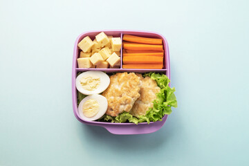 Healthy food plastic lunch container including chicken cutlets, chicken egg, salad, carrot wedges, cheese slices.