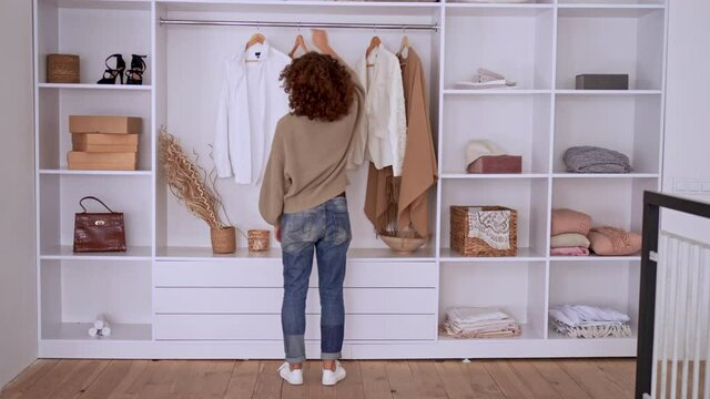 young woman with curly hair choosing outfit standing near wardrobe putting on clothes slow motion at home