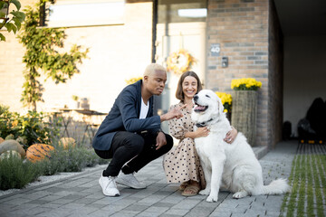 Multiracial couple caress dog on background of thier house. Concept of relationship and enjoying time together. Young smiling caucasian girl and black man. Modern lifestyle. Autumn sunny daytime