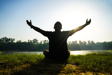 Fototapeta na wymiar silhouette of a boy doing yoga in park near sun and river - health concept