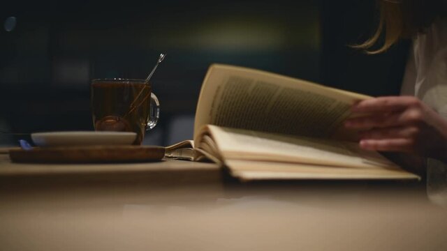 Modern female in formal outfit studies in cozy atmosphere cafe with book and cup of tea in the late evening. Education with pleasure with warm drink reading book.