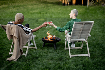 Multiracial couple drinking cocktails during home party on a green lawn in their garden. Burning fire for bbq. Concept of relationship. Black man and european girl enjoying time together - obrazy, fototapety, plakaty