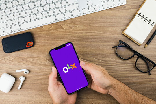 Male hand holding a smartphone with OLX app on the screen with a computer keyboard. Wooden Background. Office environment. Rio de Janeiro, RJ, Brazil. October 2021.