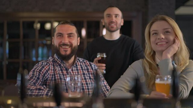 Excited Caucasian friends rejoicing goal watching sport match in pub indoors. Portrait of happy smiling men and woman talking smiling resting on weekend at bar counter with beer glasses
