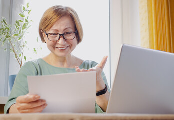 Happy middle aged elderly woman in eyeglasses reading paper document or postal letter from friend, sitting at home indoor. Reaction of pleasant paperwork bank notice or post correspondence