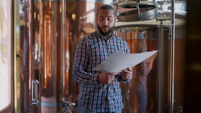Medium shot portrait of concentrated bearded brutal male brewer analyzing paperwork standing in brewery at large beer tanks. Handsome confident Caucasian man checking quality of production process