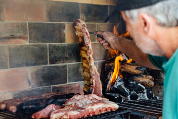 Asado. Argentinian mature man making a barbecue in the grill of his house. He is prodding the roasted meat with a fork. - obrazy, fototapety, plakaty