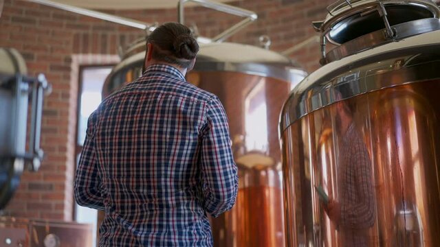 Serious professional brewer with tablet turning checking high-quality beer production indoors. Happy satisfied Caucasian bearded man controls alcohol manufacturing process in brewery
