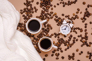 Two cups of black coffee, alarm clock, white sweater and coffee beans on a beige background. Cozy date and wellness. Top view