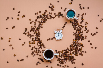 Two cups of black coffee in a circle from coffee beans and a white alarm clock on a beige background. Time to drink coffee concept. Top view