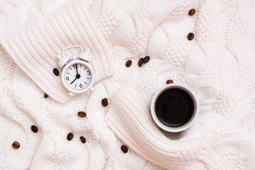 Cup of black coffee, alarm clock and coffee beans on a cozy white sweater. Time to drink coffee concept. Top view