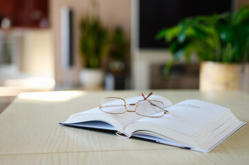 Open book with glasses on a light wooden table against the background of the room. Selective focus...