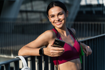 Fototapeta na wymiar Healthy brunette woman holding smartphone and smiling widely to the camera after hard training at the stadium. Sport concept